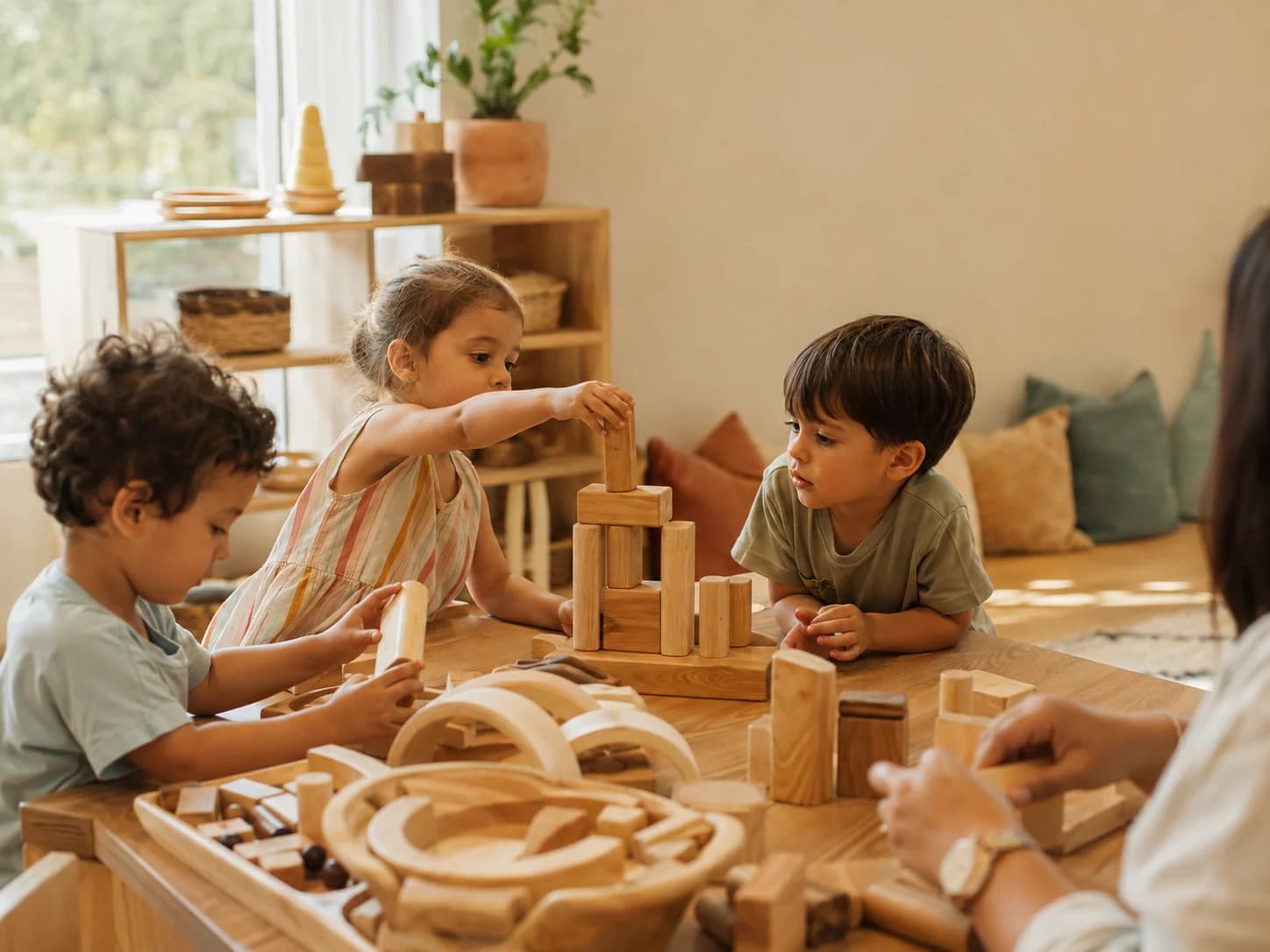 Young children building a tower from natural wooden blocks at a low table in a bright, warm early-years room