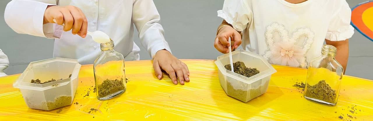 Children's hands at a yellow table in a Kids Are Kids classroom, spooning sand and soil into small glass jars